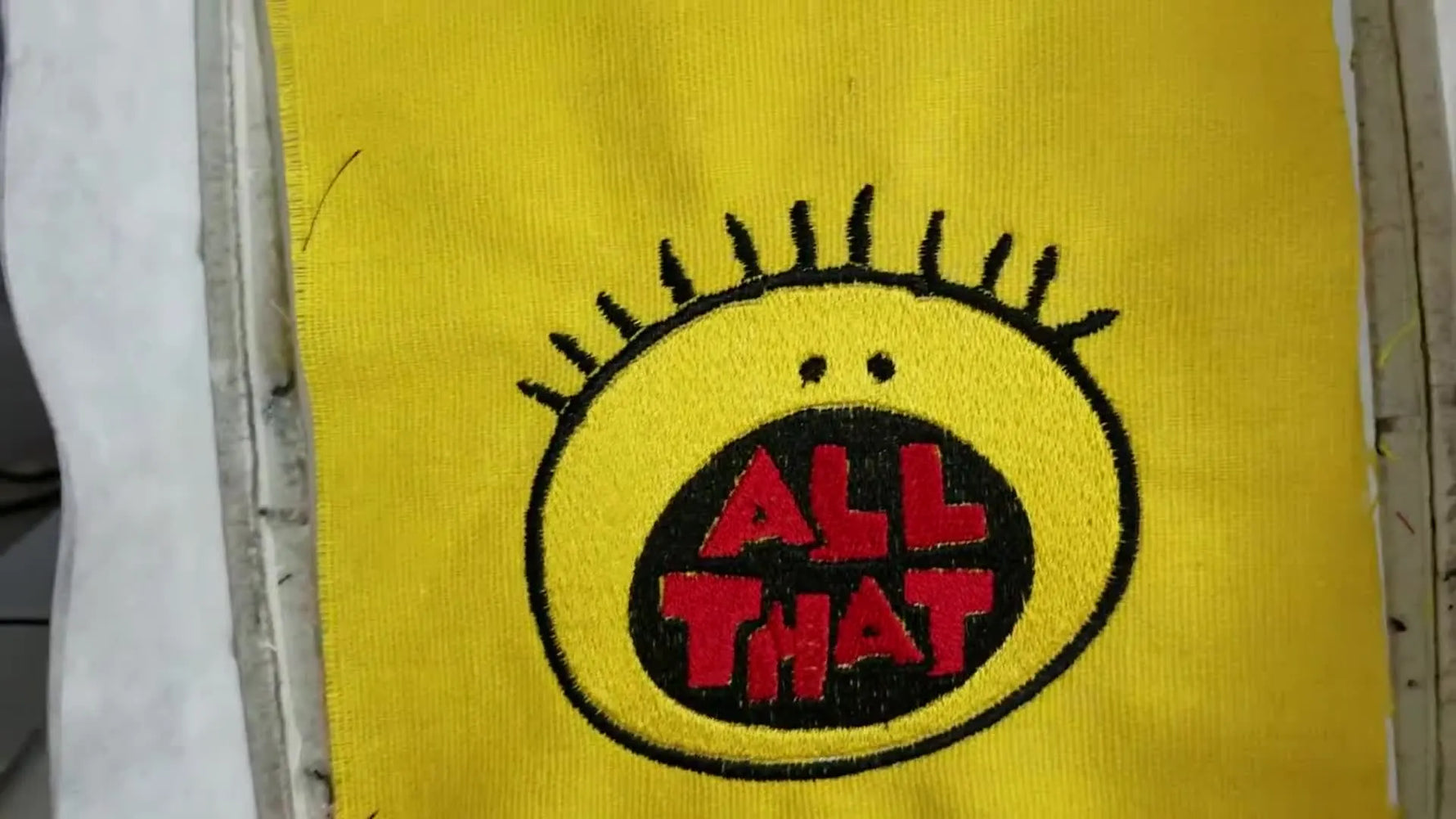 A finished “All That” logo patch sits cleanly in the hoop, showing crisp red lettering and a black satin border over a yellow fill.