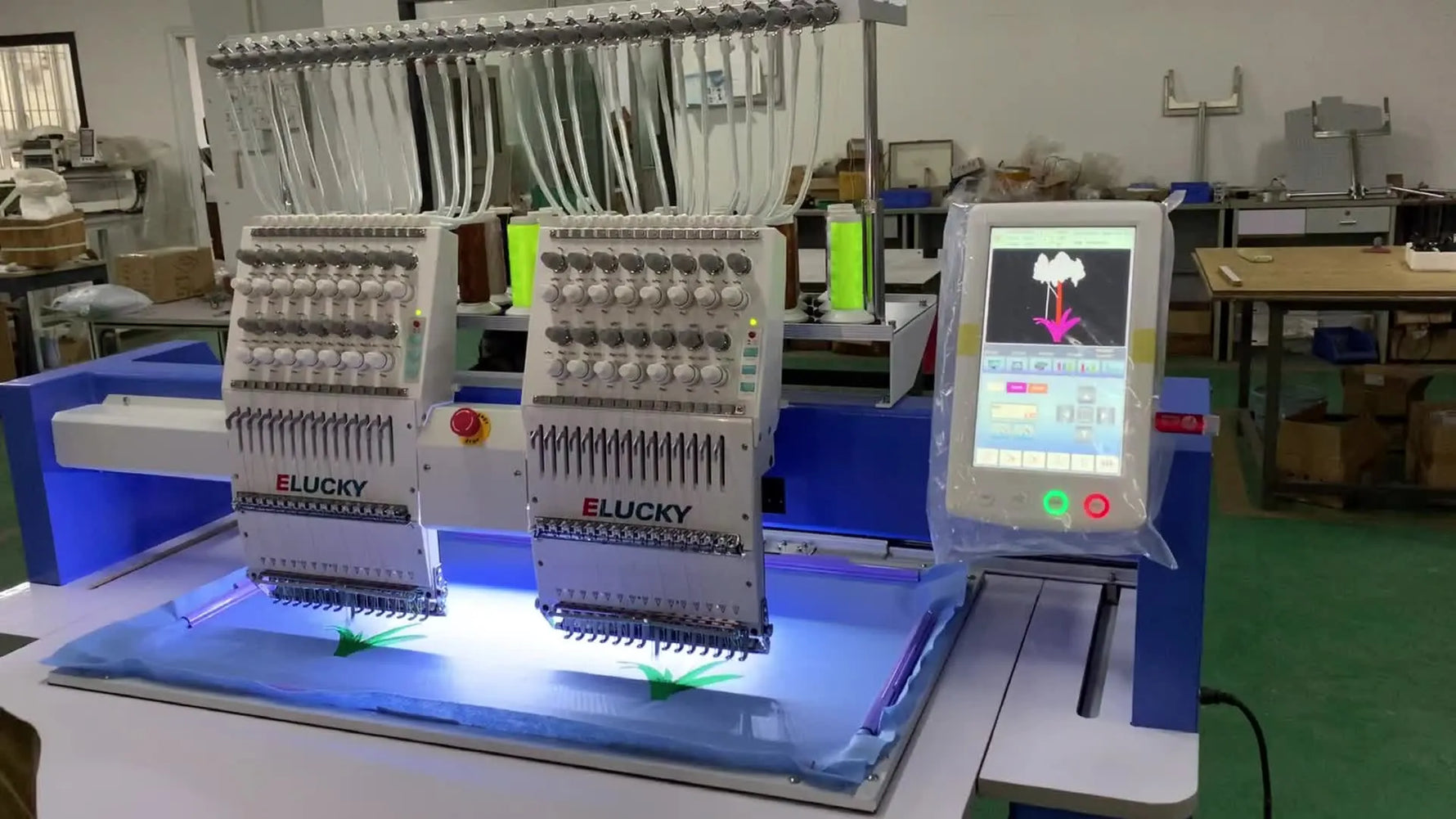 Two heads of an ELUCKY commercial embroidery machine stitch a multi-color flower design on a large blue hooped fabric sheet.