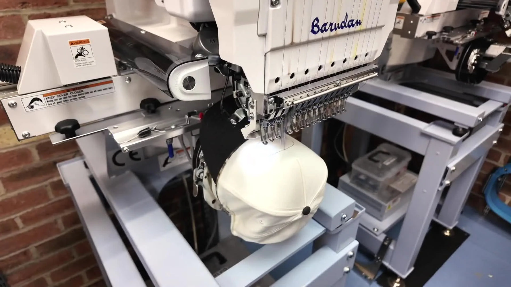 A close-up shows hands aligning a woven patch onto a trucker hat while it’s mounted on a Barudan cap driver, right after the placement trace stitch.