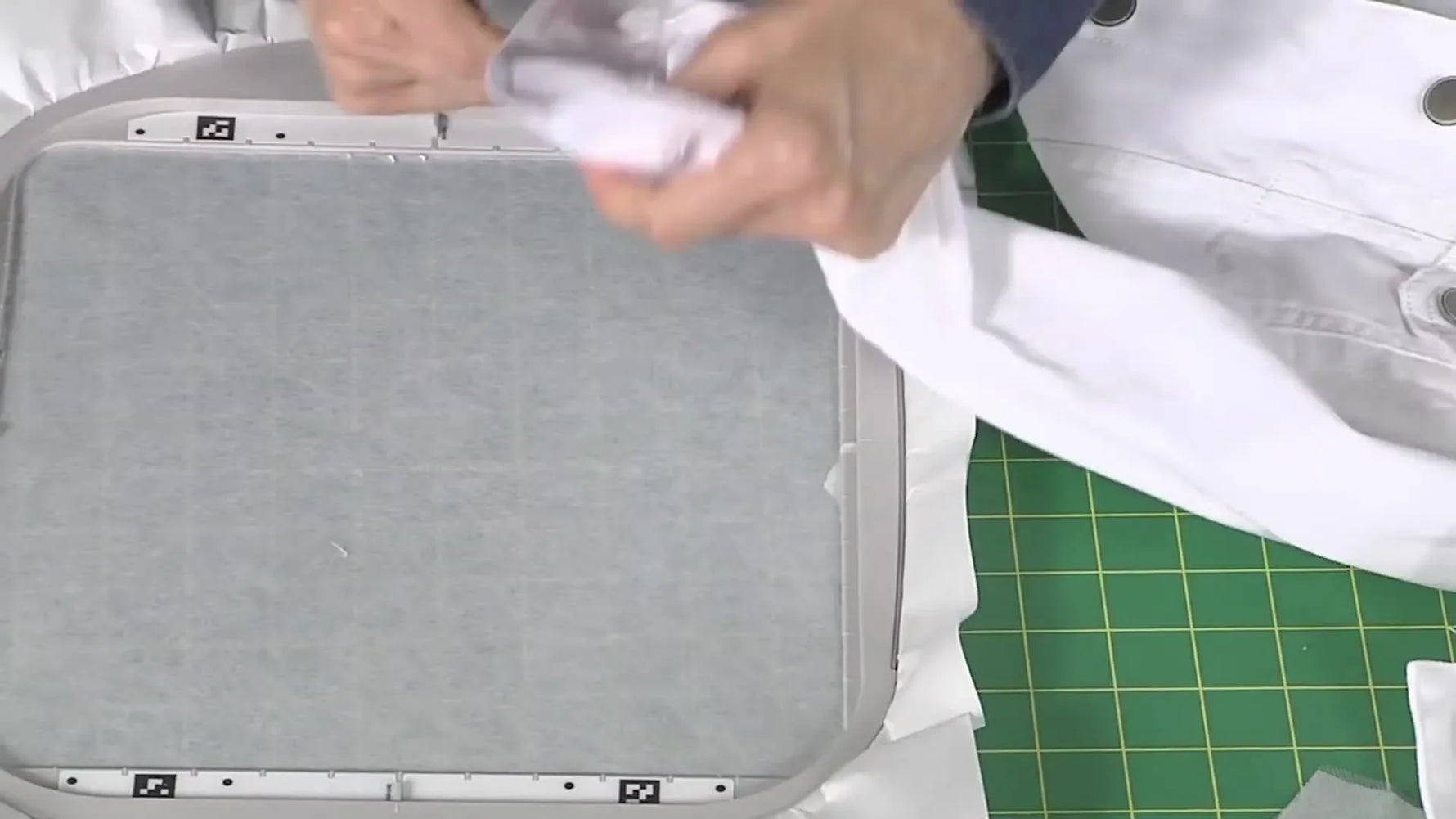 A smartphone is held parallel over a hooped sleeve cuff while an embroidery positioning app captures the hoop and marker for precise design placement.