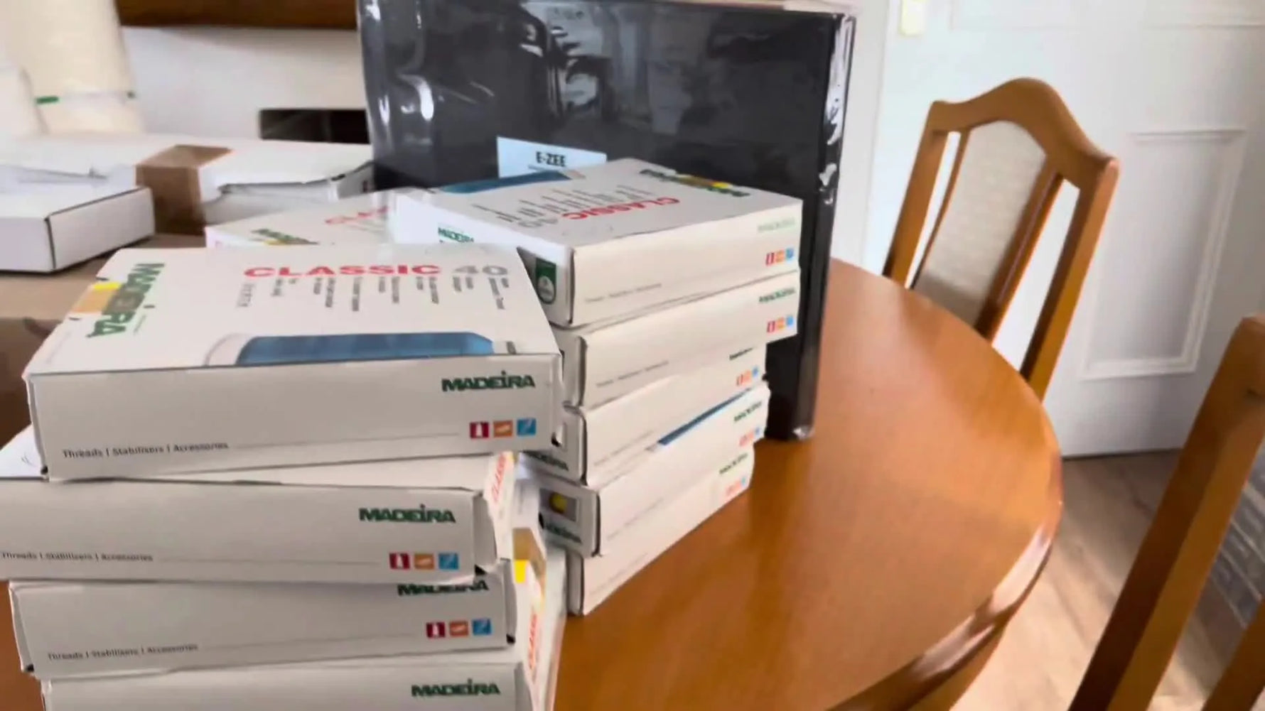 A wide view of a kitchen table fully covered with Madeira thread boxes, stabilizer, foam, and bobbin supplies before being organized into working inventory.