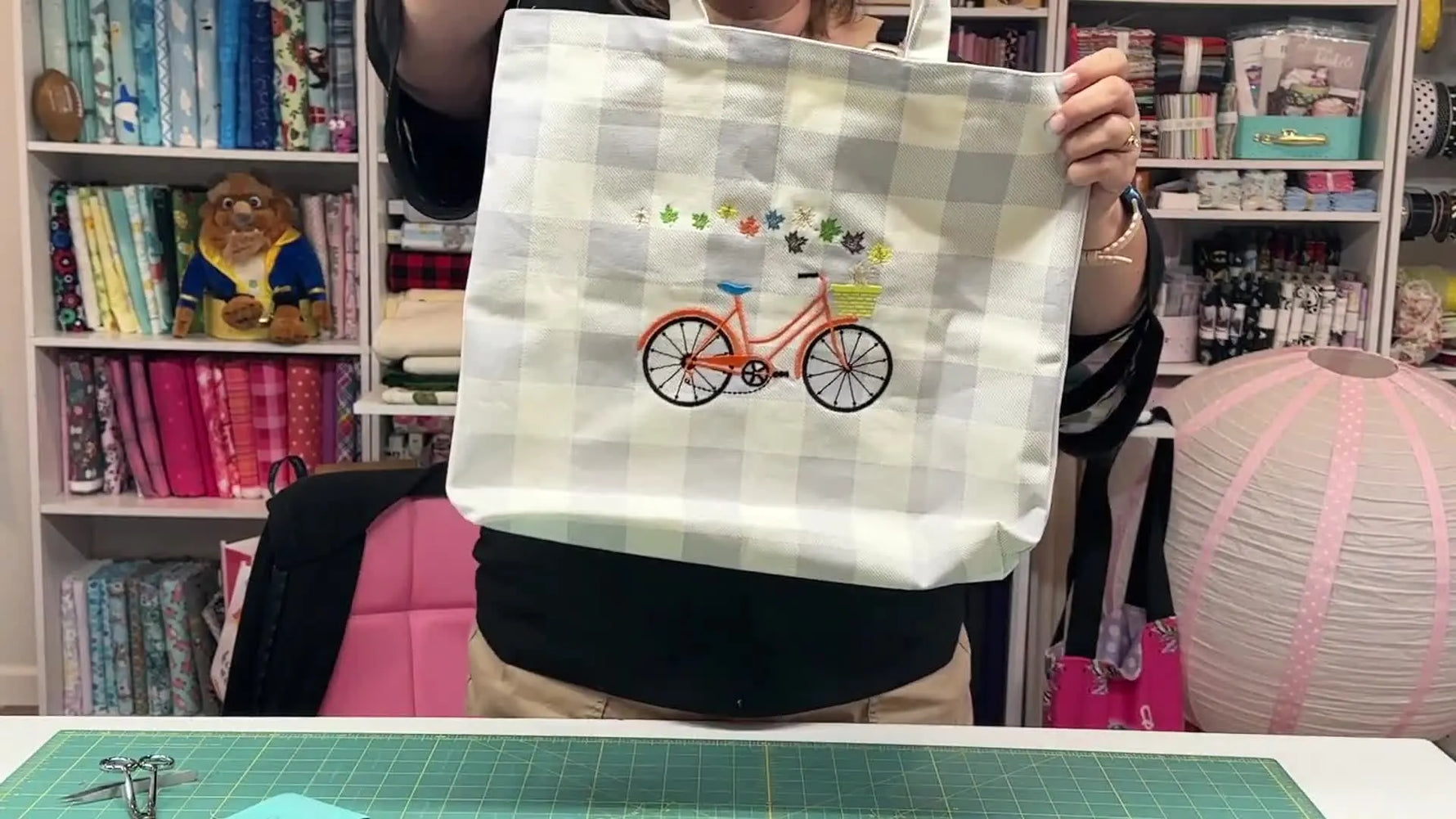 The instructor holds up the finished Kimberbell buffalo check tote with a colorful bicycle embroidery stitched cleanly in the center.