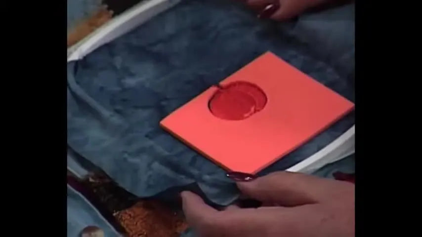 A close-up of an embroidery hoop showing sticky stabilizer in the hoop, denim floated on top, and a square of orange Puffy Foam positioned in the stitch area.