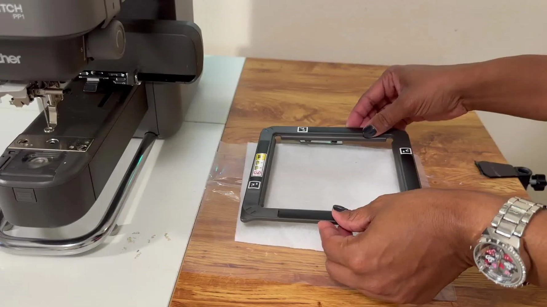 Hands snap the Brother Skitch 4x4 magnetic frame closed over felt and clear plastic sheeting, showing the fast, screw-free hooping method used for the patch.