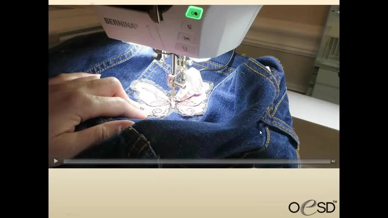 Close-up of hands guiding a denim pocket with a butterfly patch under the sewing machine needle while tacking it in place.