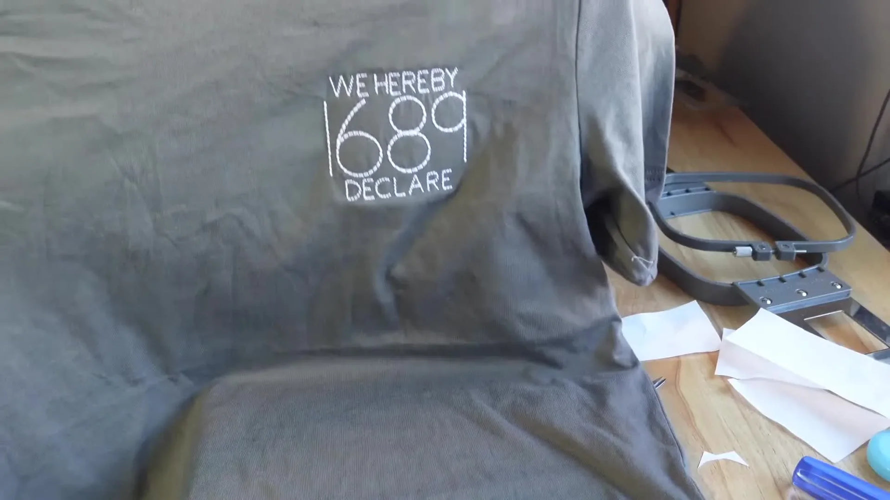 A finished grey Bella+Canvas t-shirt is held up to show a neatly centered left-chest embroidery result.