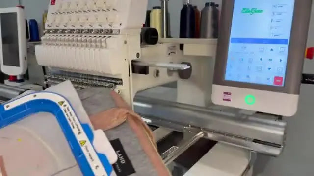 A heavy diaper bag pocket is being slid onto a multi-needle embroidery machine arm while the operator supports the bag’s weight to prevent drag and misalignment.