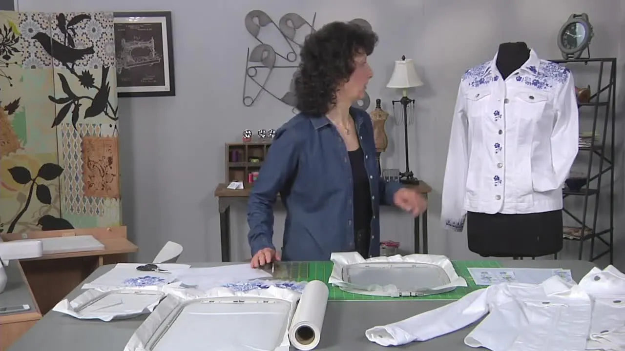 A white jean jacket on a dress form featuring blue floral embroidery on the shoulders, yoke, and sleeve cuff.