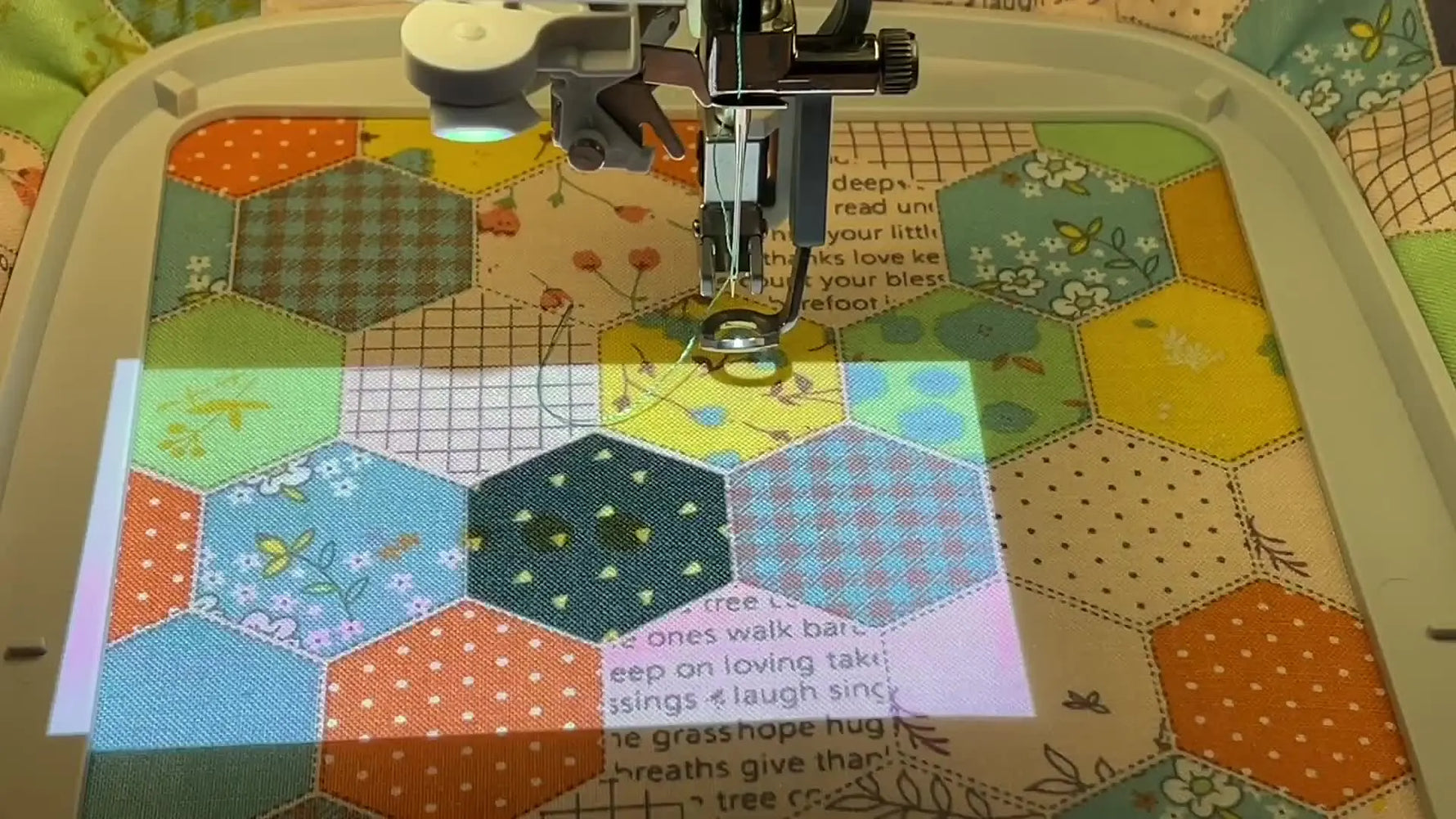 A flower embroidery design is projected onto a hooped hexagon patchwork block, showing precise on-fabric placement before stitching.