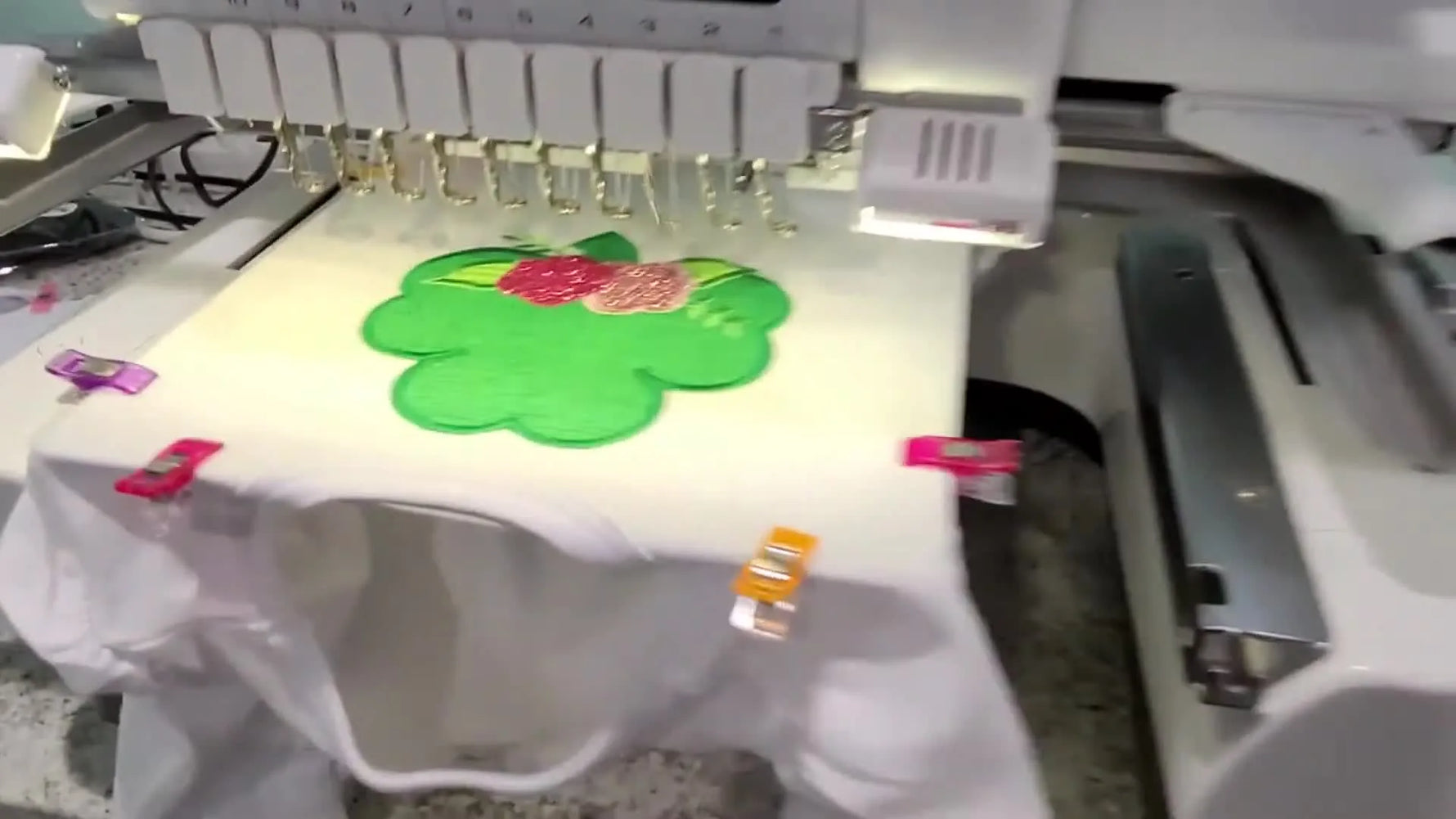 A close-up view of a multi-needle machine stitching a satin border around pink glitter HTV flowers on an appliqué design.