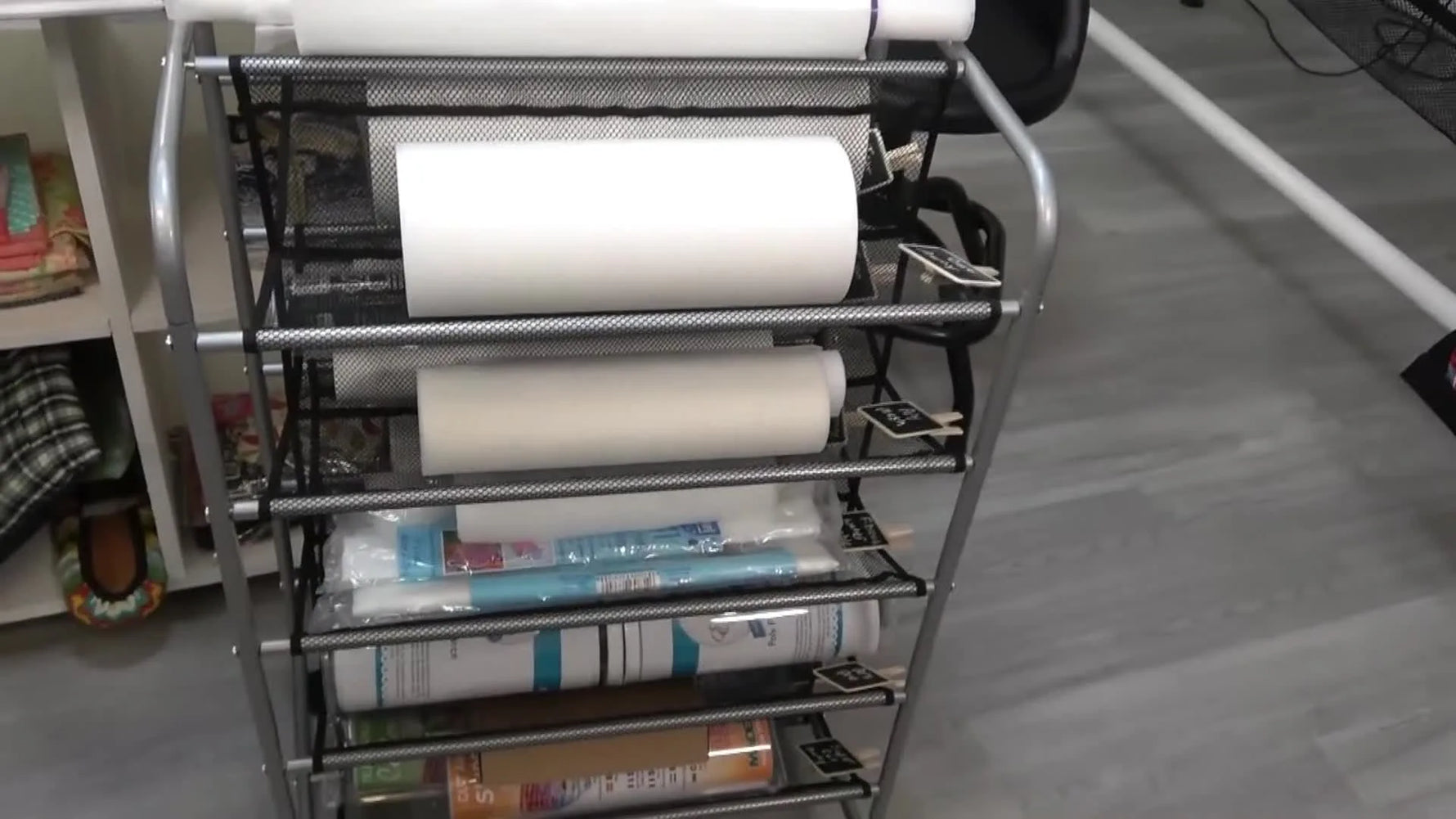 A five-tier rolling mesh shoe rack packed with embroidery stabilizer rolls and labeled with clip-on chalkboard tags.