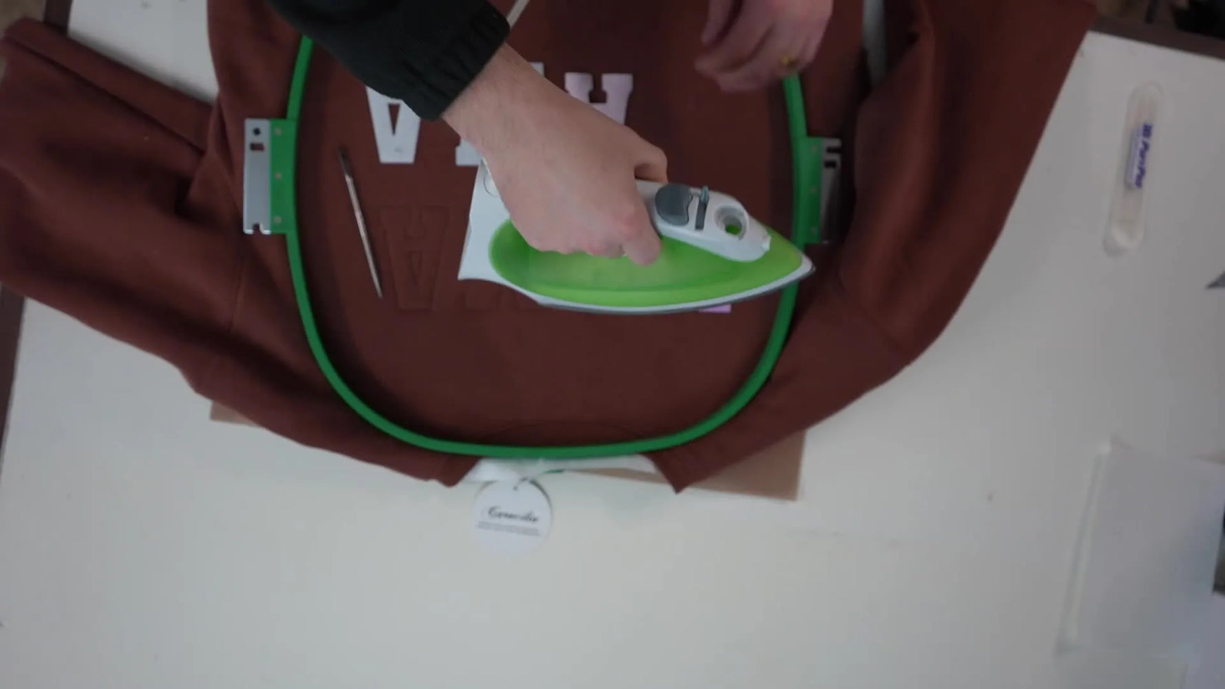An overhead shot of an iron fusing appliqué letters directly onto a hooped sweatshirt before the final satin border run.