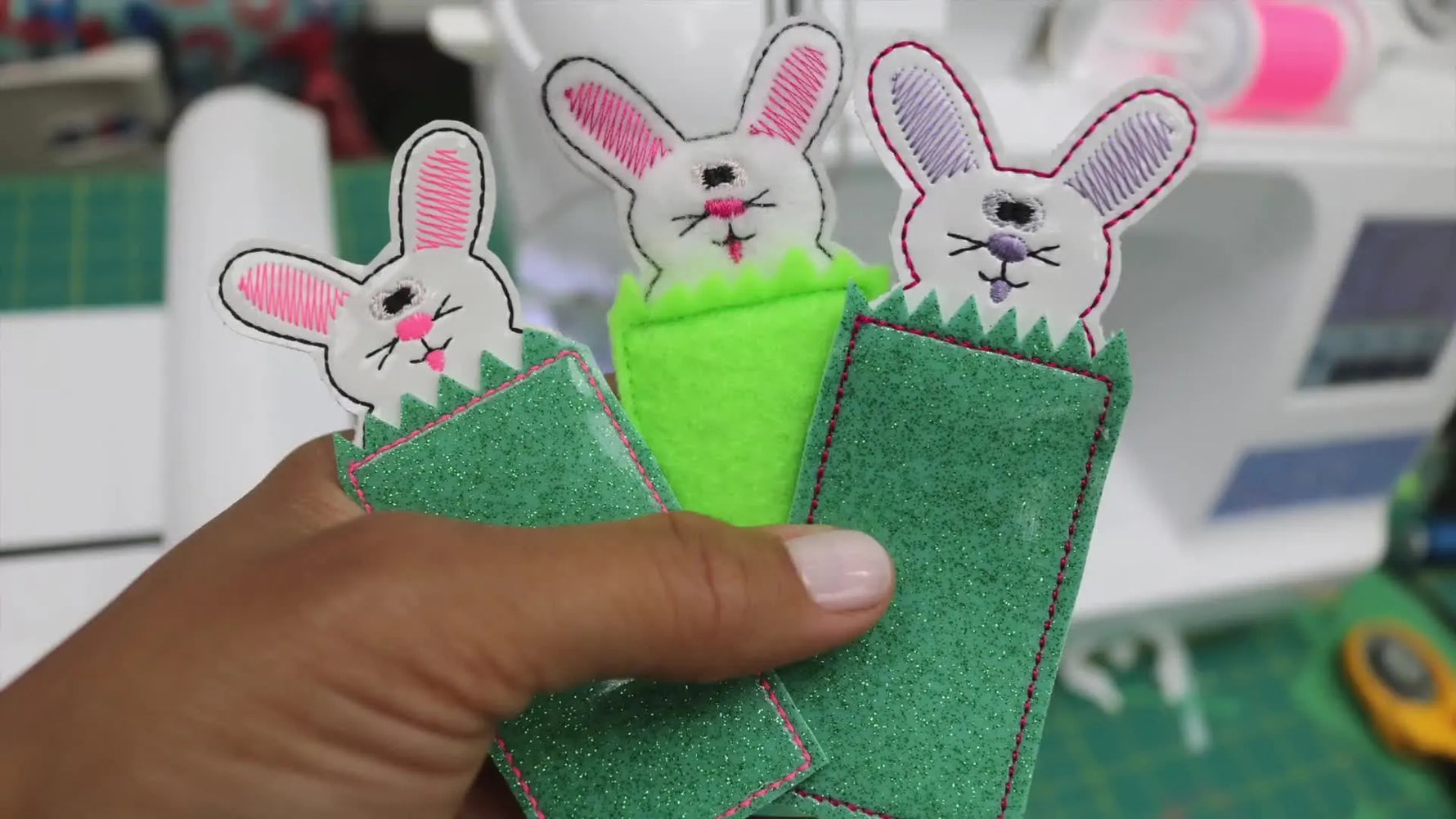 Three finished ITH bunny bookmarks in bright vinyl colors are held above a cutting mat, showing the clean two-sided construction and grass detail.