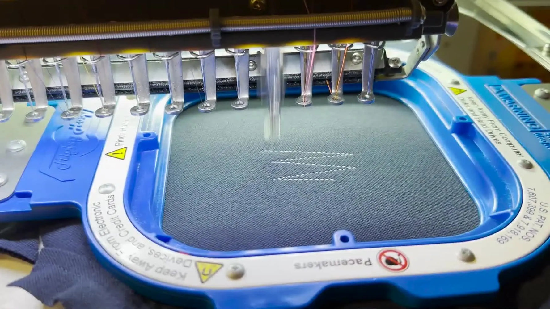 A multi-needle embroidery machine begins stitching on dark fabric held tight in a blue magnetic hoop, setting up a high-contrast sample run.
