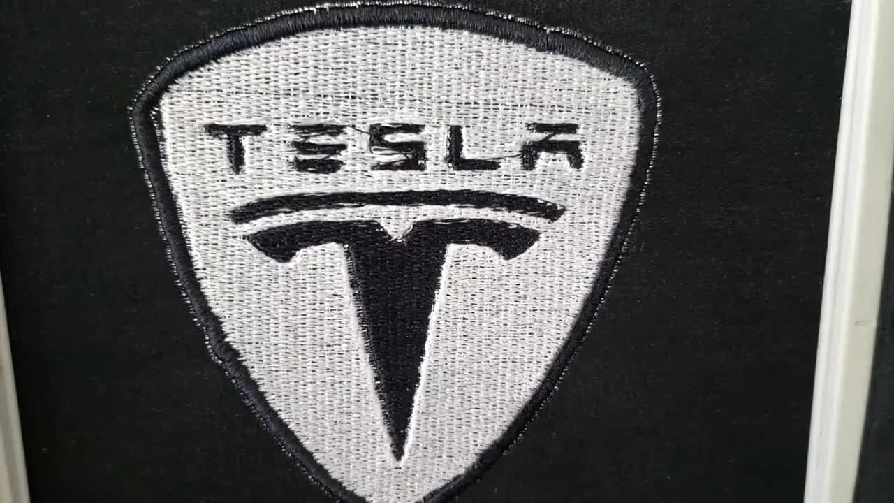 A neatly cut Tesla logo patch is held in hand, showing clean satin edges ready to be turned into a magnet.