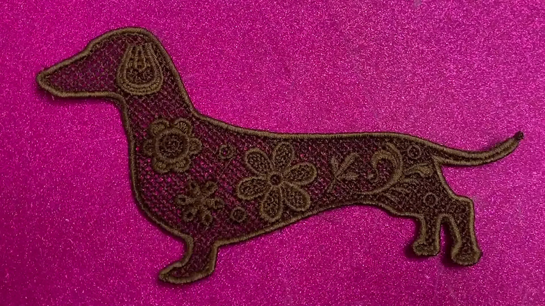 A close-up of the finished freestanding lace dachshund patch stitched in brown on a pink background.