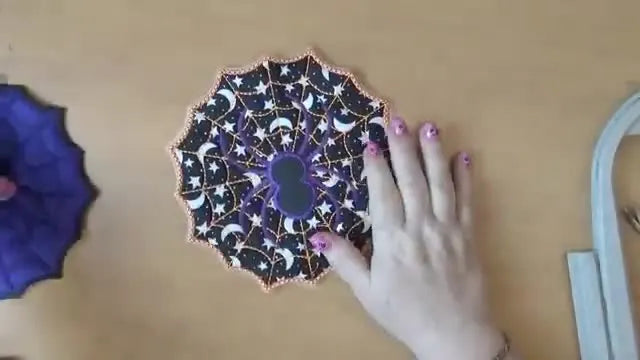 Two finished spider coasters in bright orange and purple thread are shown side by side, highlighting the bold web stitches and the two-tone border trick.