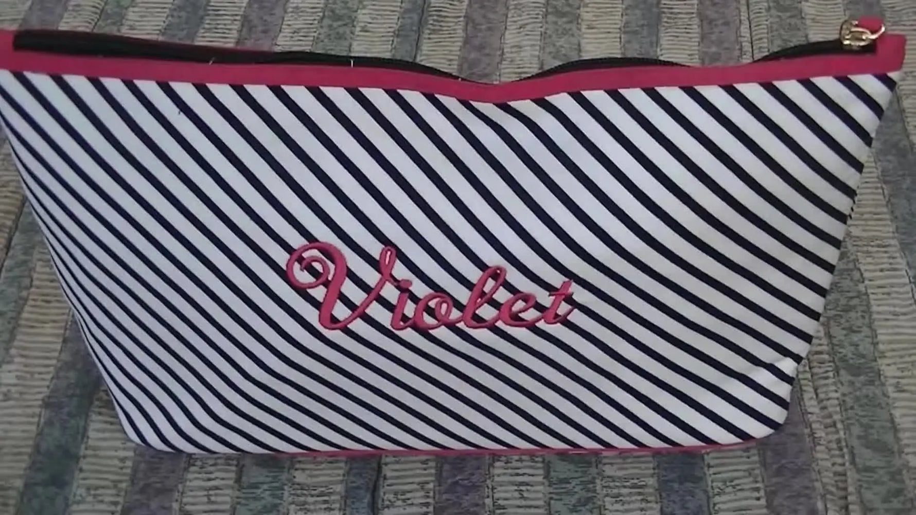 A finished striped cosmetic bag stands upright with a pink zipper trim and matching pink “Violet” script embroidery centered on the front.