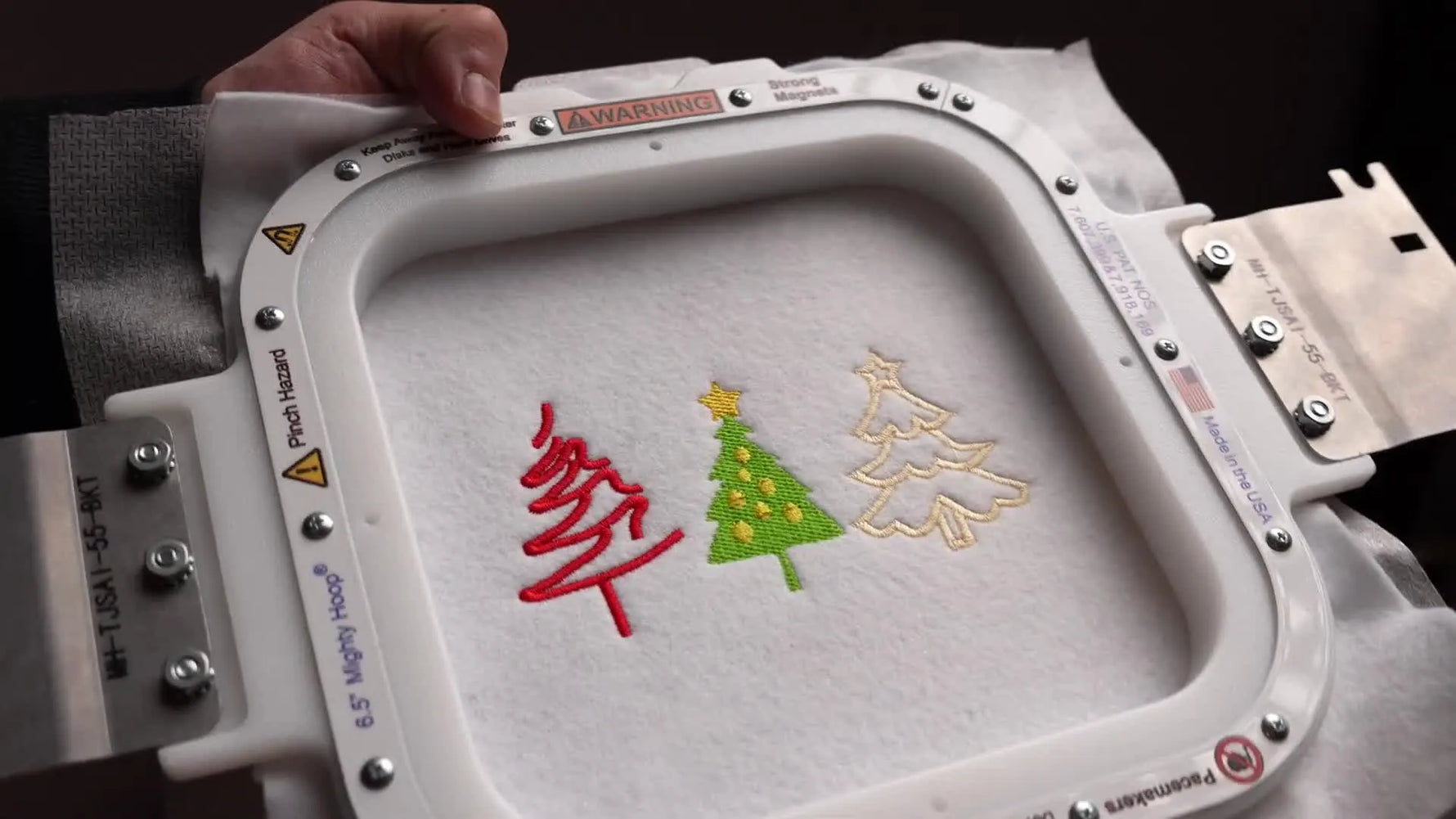 Overhead view of a square magnetic hoop holding felt with three finished Christmas tree embroidery designs stitched out on a Tajima machine.
