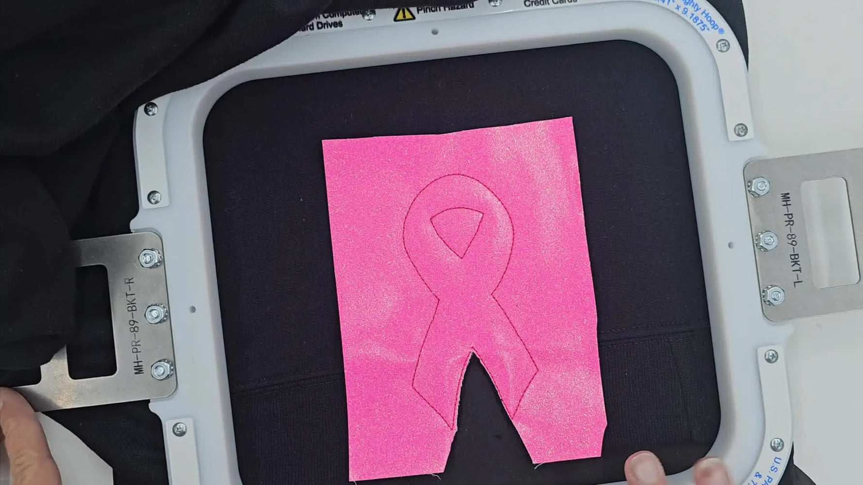 A fully trimmed sparkly pink ribbon appliqué sits in a magnetic hoop on a black sweatshirt sleeve, ready for the final satin stitch.