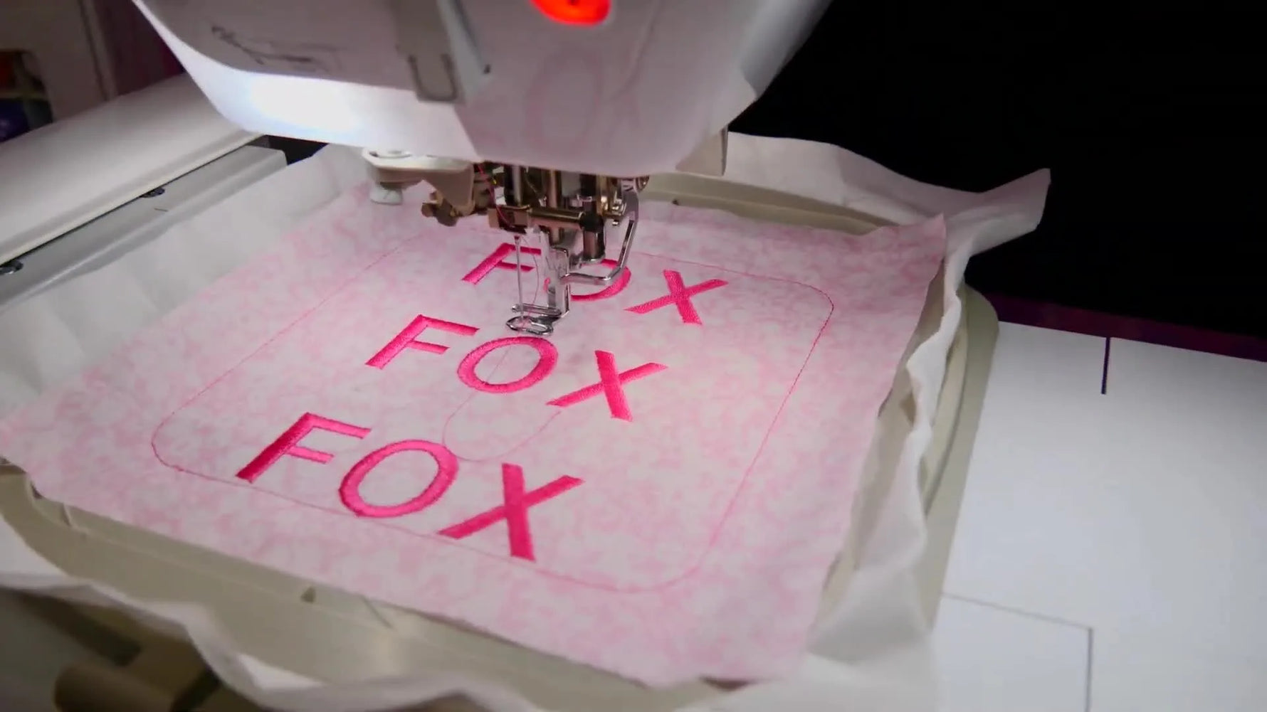A close-up of the embroidery machine stitching a pink “FOX” design in the hoop, showing clean needle penetration and stable fabric support.