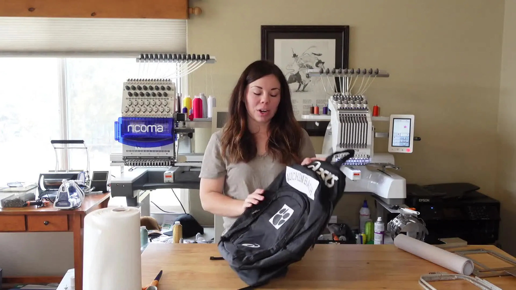 Woman holding up a black Asics backpack with two white patches on the front pocket: one with the name 'AUCHENBACH' and another with a paw print outline.