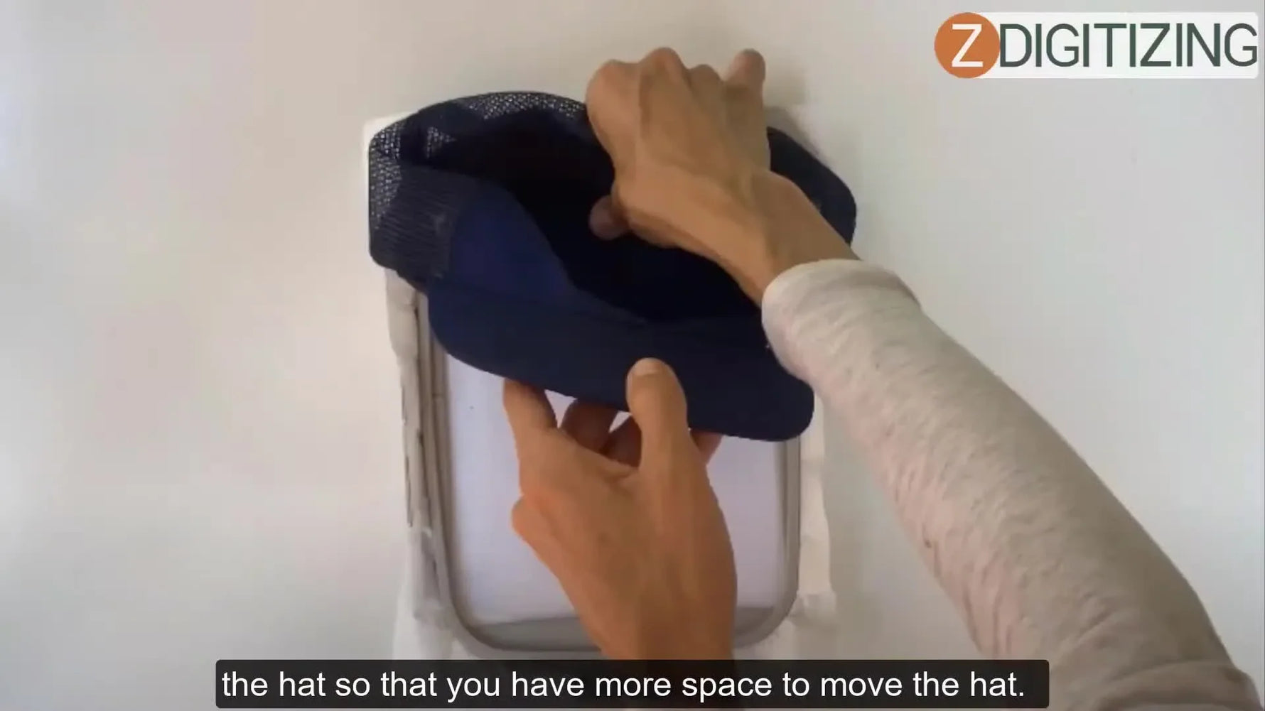Hands press the crown of a blue mesh cap onto a hooped sheet of sticky stabilizer, demonstrating the floating method on a flat hoop.