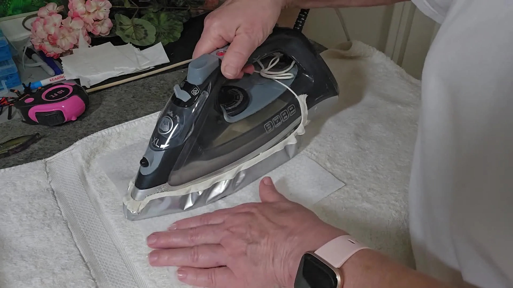 An iron begins a gentle center-out press on the back of an embroidered block laid face-down on a towel.