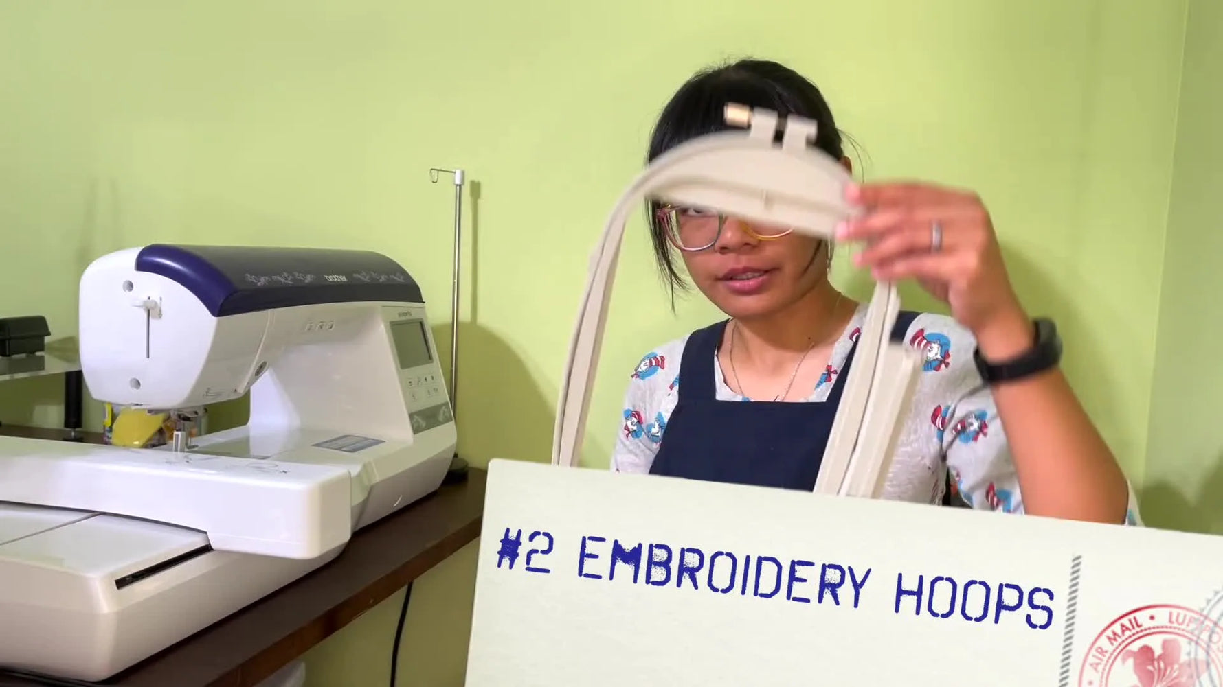 The host holds up a 6x10 embroidery hoop next to a flatbed Brother machine to show the real-world scale of a larger hoop size.