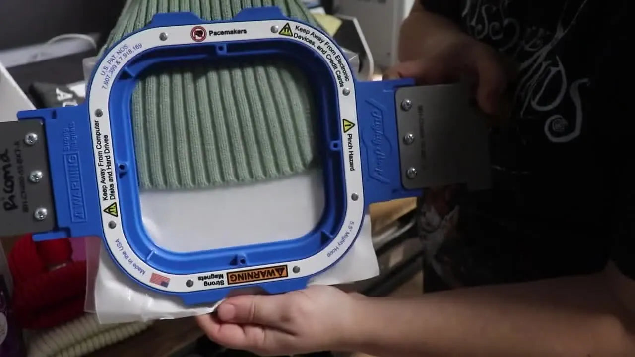 A sage ribbed knit beanie is held taut inside a blue 5.5-inch magnetic hoop, showing the clean, even hooping tension needed for crisp faux chenille lettering.