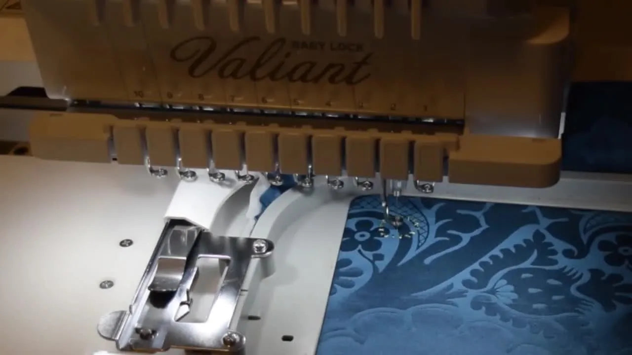 A close-up of a multi-needle embroidery machine stitching a pink floral center onto dark blue fabric inside a hoop.