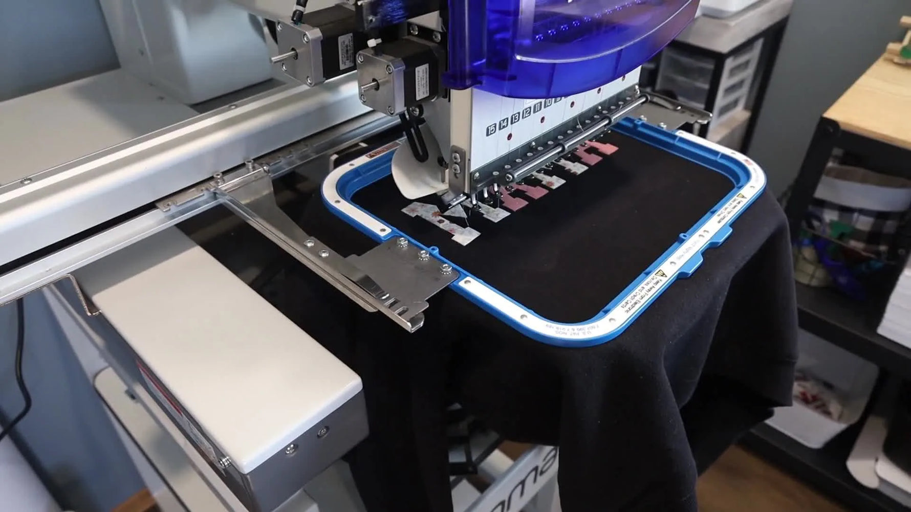 A close-up of a multi-needle embroidery machine finishing a satin stitch border around the floral “MAMA” appliqué on a black sweatshirt.