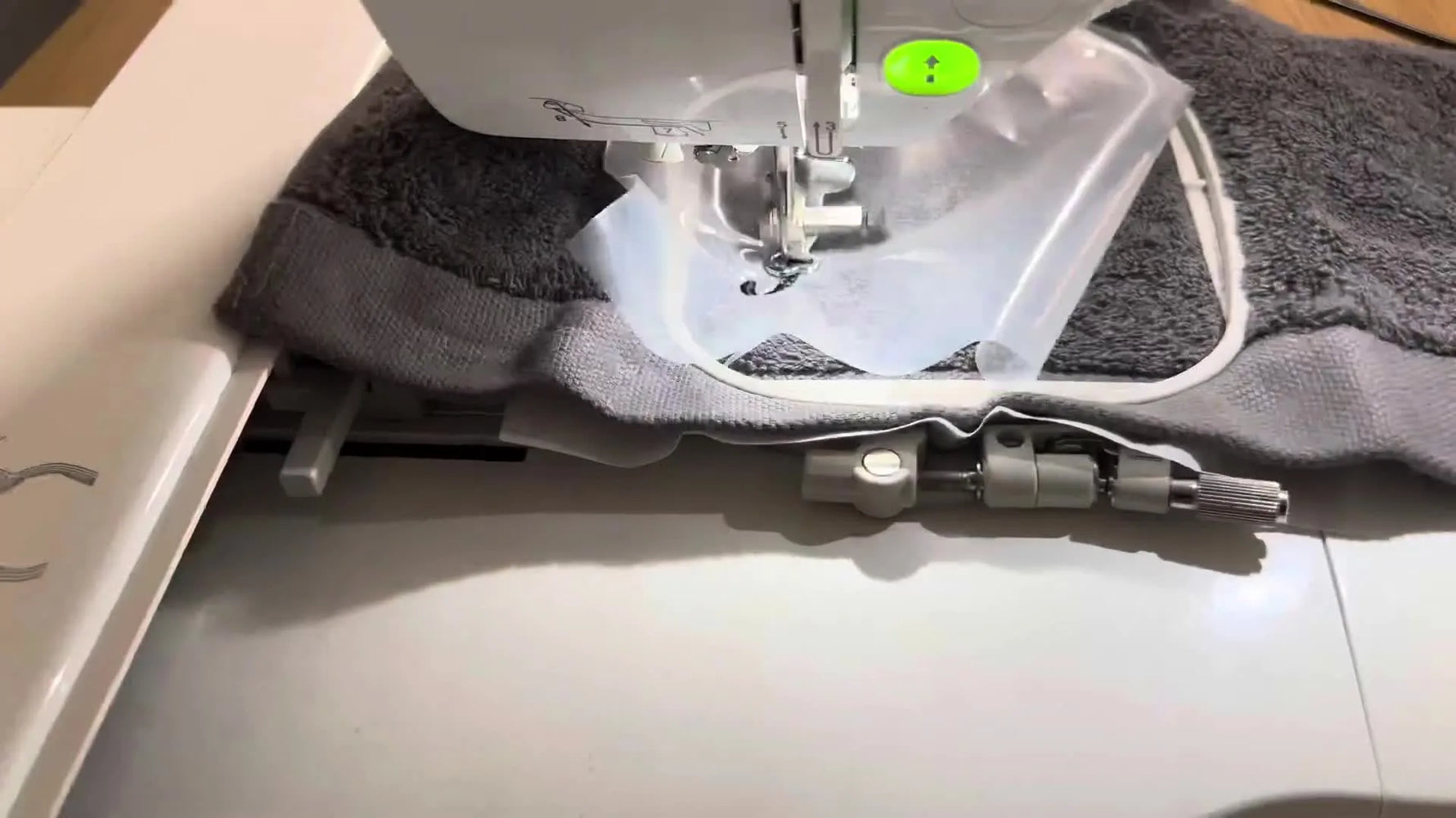 A close-up of the Brother NV870SE stitching a name through clear water-soluble topping on a thick charcoal towel.