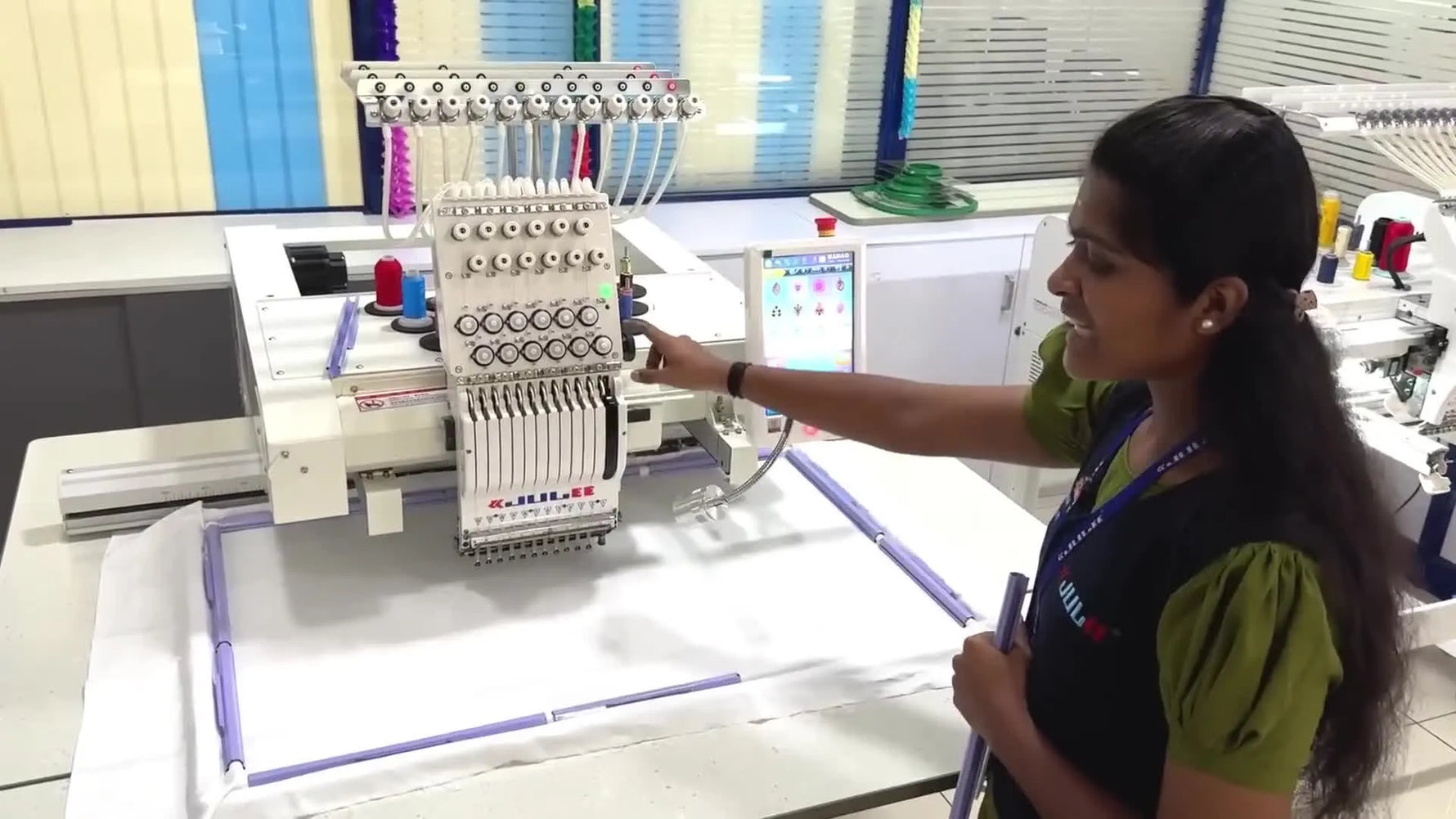 A wide view of the JUILEE single-head 12-needle embroidery machine with the operator pointing at the head and thread setup.
