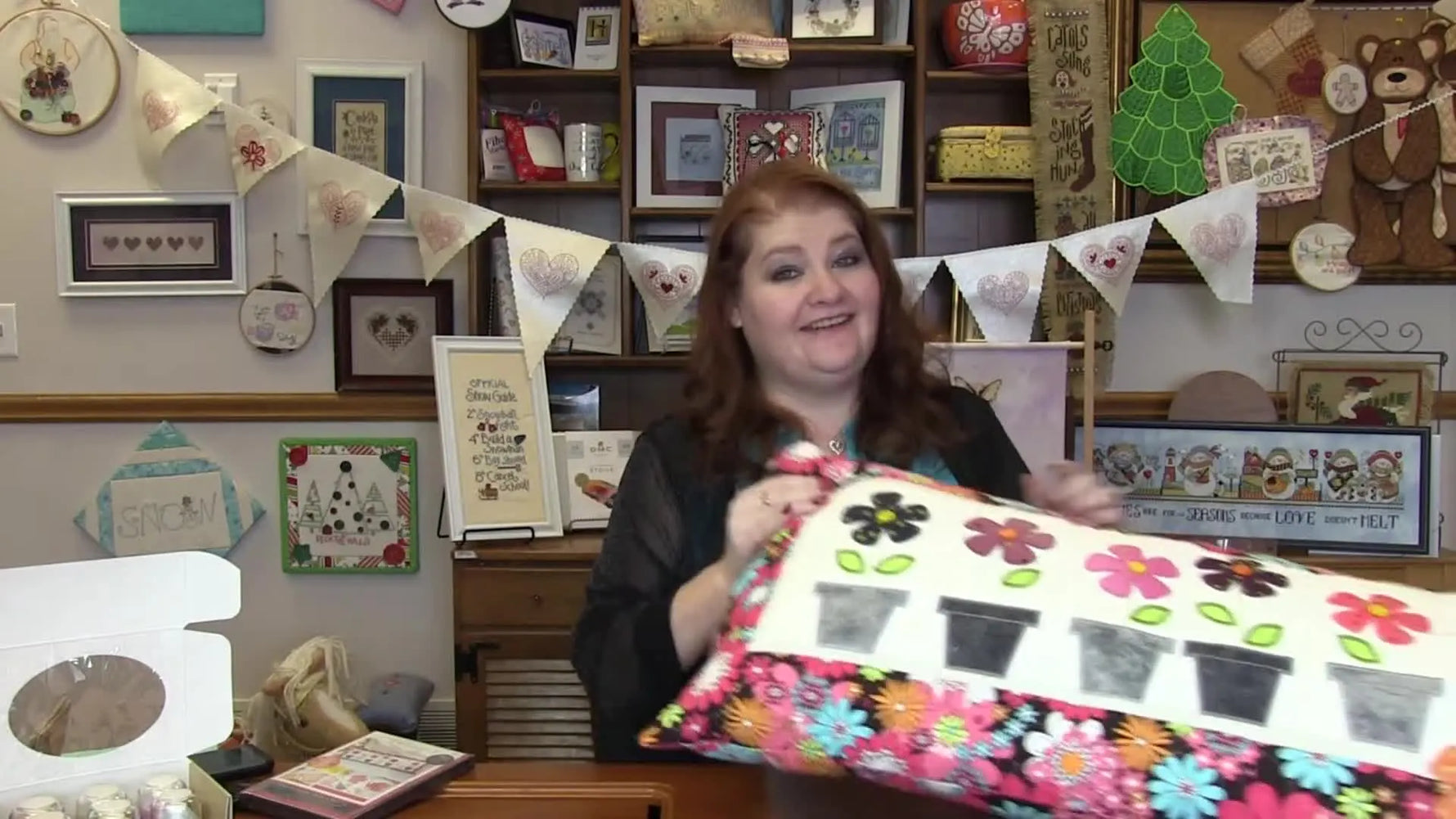 The host holds up the finished Kimberbell “May Flowers” bench pillow, showing the row of potted appliqué flowers and stitched details.