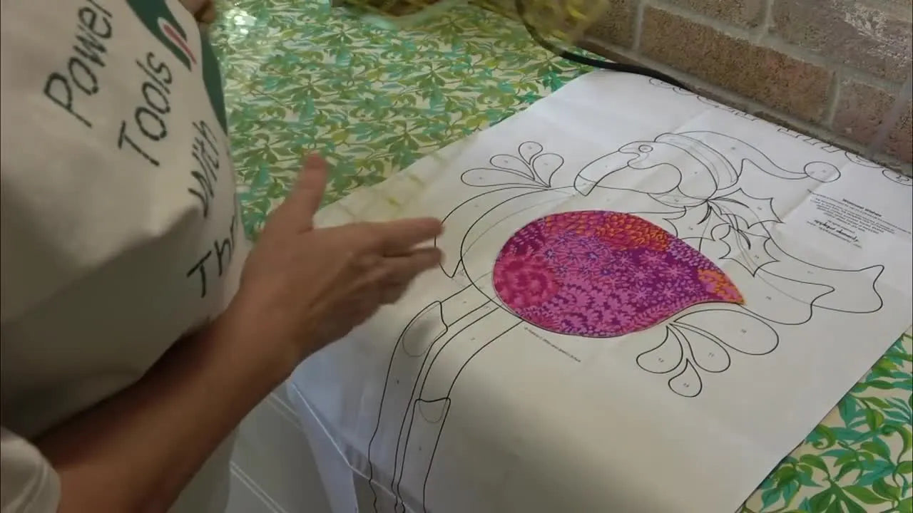 Hands measure a paper appliqué pattern with a large square quilting ruler to confirm angles and overlap before splitting the design into multiple hoopings.