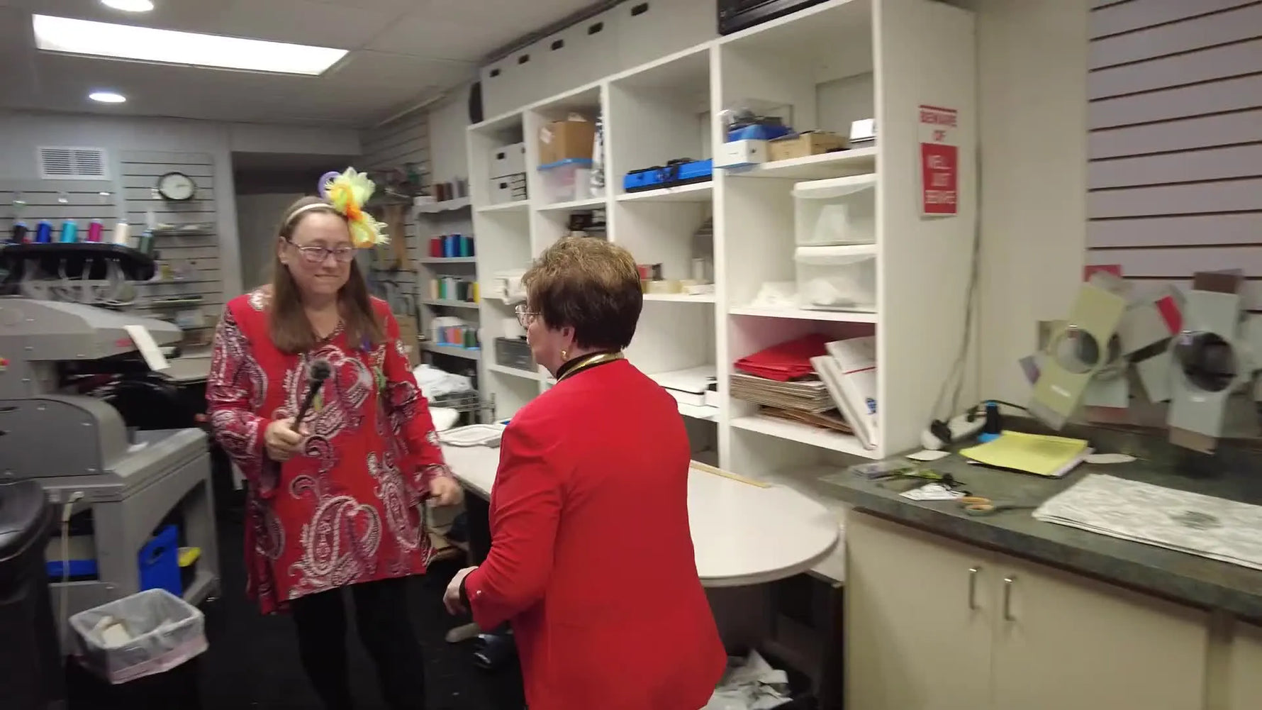 Miss Dell and Julia walk toward the Melco machines in a working commercial embroidery shop, with thread and tools visible in the background.