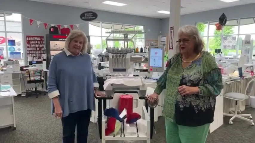 Two presenters stand beside a Brother PR1055X 10-needle embroidery machine in a retail classroom setting, highlighting the machine as the centerpiece of a side-hustle training event.