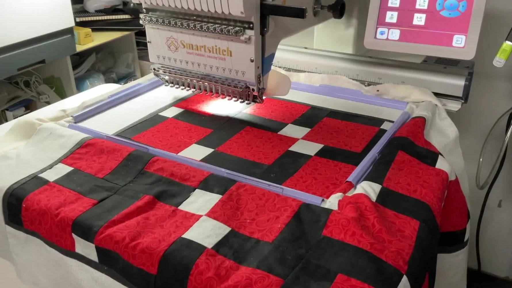 A red-and-black patchwork quilt sandwich is clamped flat inside a large rectangular magnetic/aluminum frame on the SmartStitch machine bed.