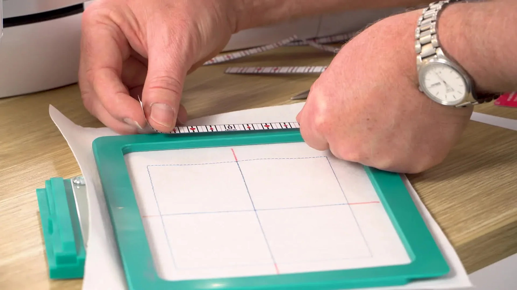 An extreme close-up shows the adhesive centering ruler being aligned precisely to a red pen mark on the magnetic hoop frame.