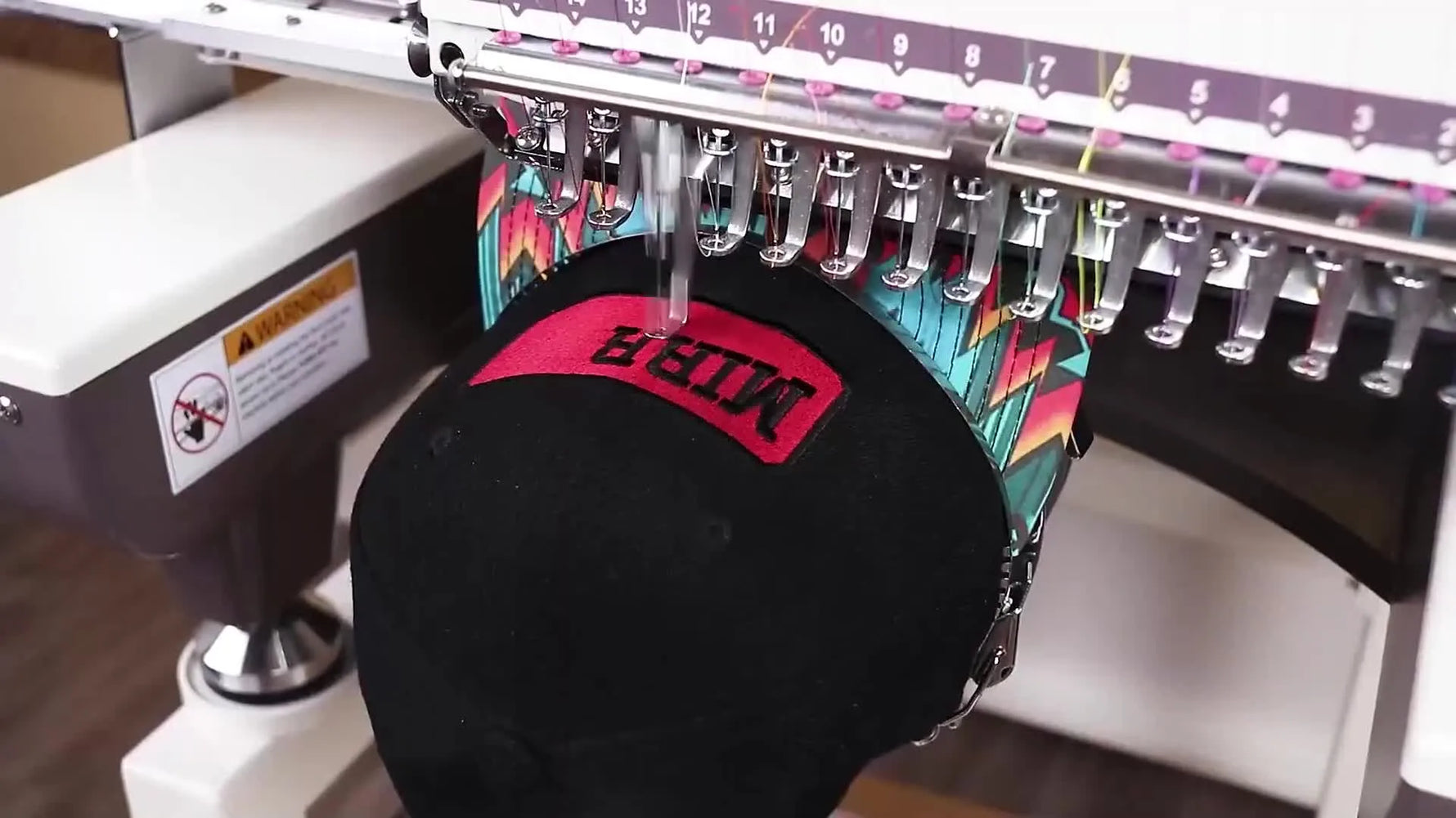 A commercial multi-needle machine stitches a logo onto a black baseball cap on a cap frame, showing the real-world setup that drives hat-production speed.
