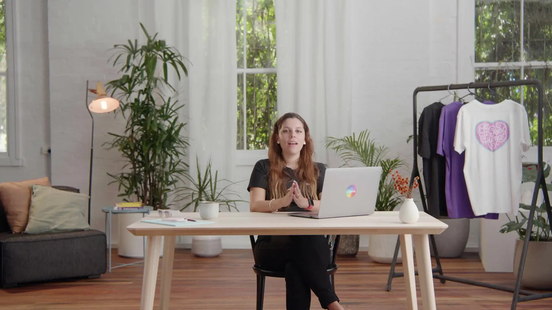 Woman sitting at a desk with a laptop, gesturing towards a clothing rack with various graphic t-shirts behind her.