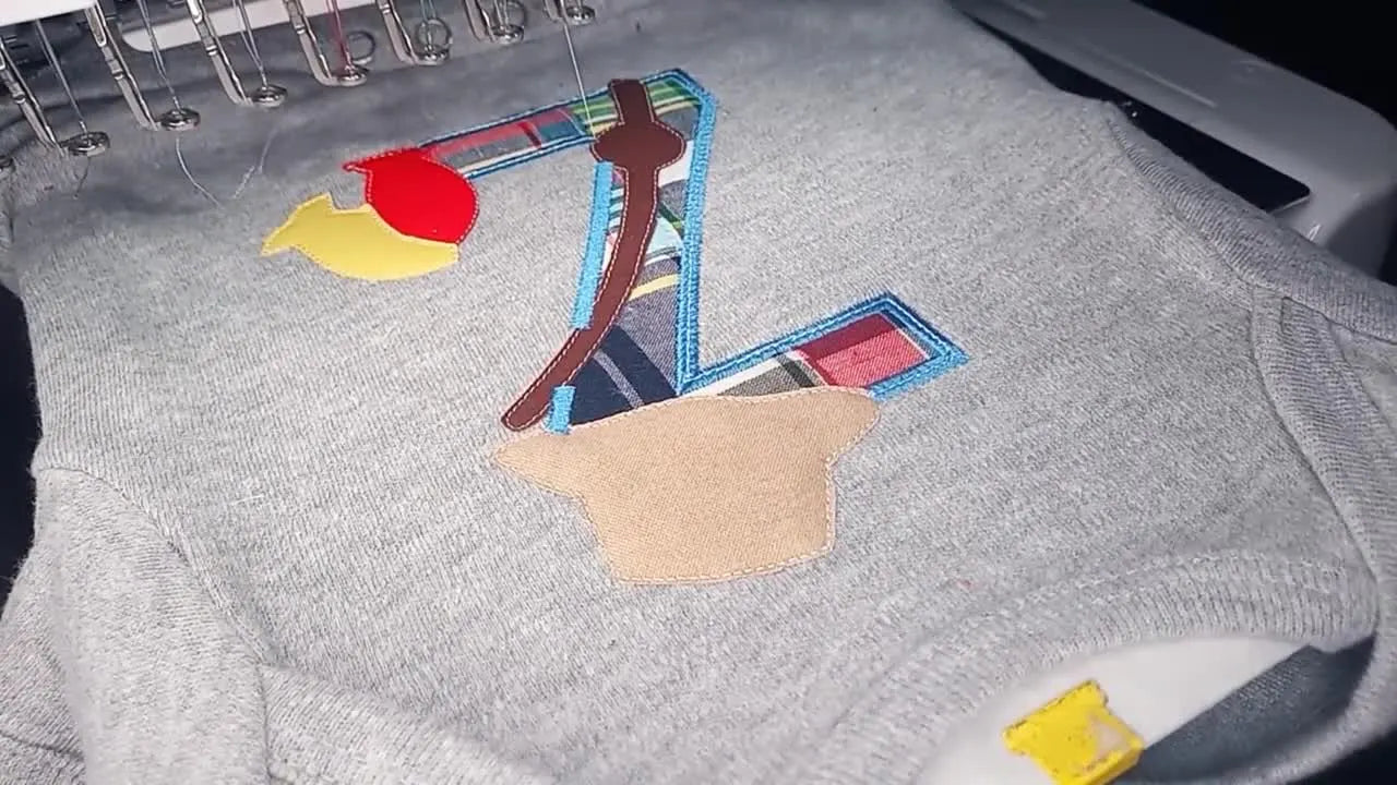Close-up of the Brother PR1000e stitching a dense satin border over a raw-edge appliqué letter on a hooped baby onesie.