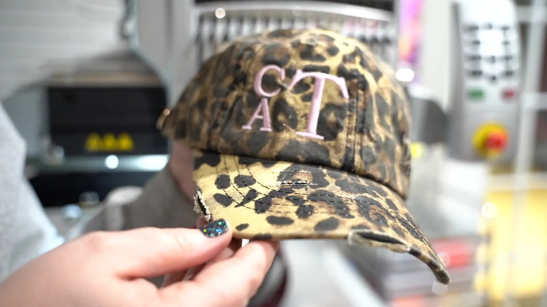 A person's hands holding up a leopard print baseball cap with pink embroidered initials 'CAT' on the front.