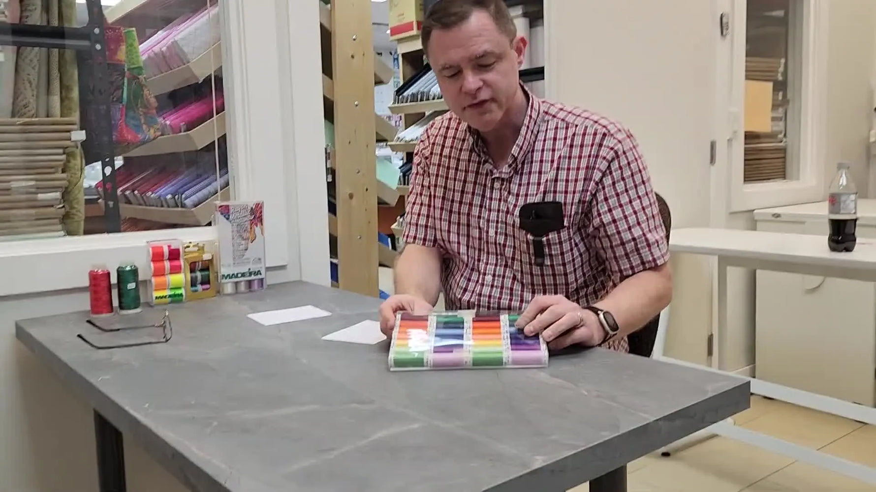 The host holds up a 28-spool Mettler Poly Sheen box, showing a rainbow gradient of colors in a clear case.