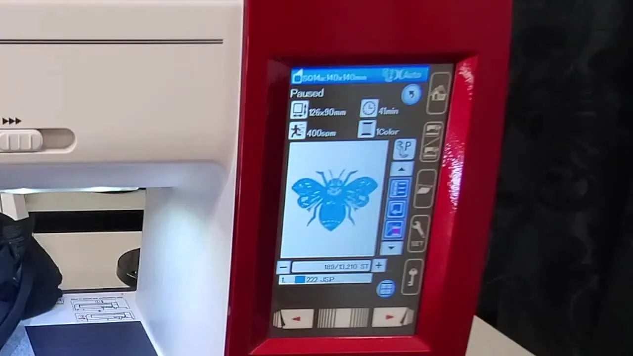 A close-up of the embroidery machine stitching a white monochrome bee design onto a black knit T-shirt inside the hoop.