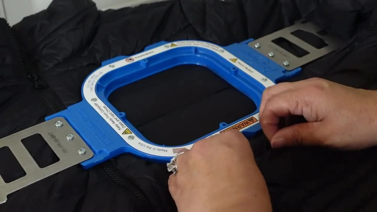 Hands pull the puffy jacket’s outer shell away from a 5.5-inch magnetic hoop to flatten the dome and create a tight, stitch-ready surface.
