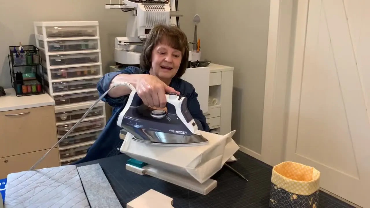 A close-up shows a steam iron pressing firmly on a tote bag’s boxed corner over the Totally Tubular Pressing Station for a sharp, professional crease.