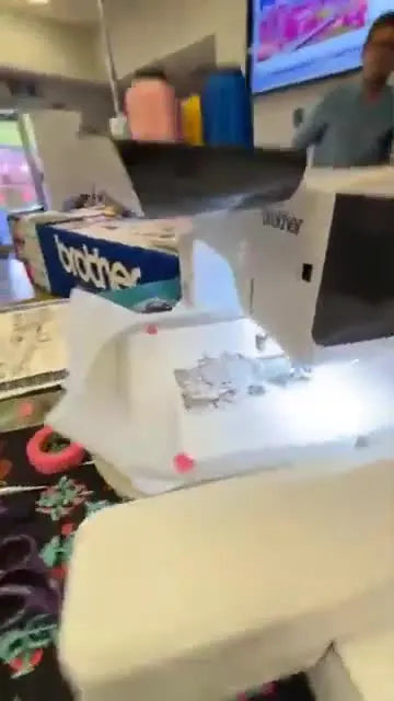 A close-up of a Brother embroidery machine stitching black run-stitch line art onto watercolor paper secured with pink tape in the hoop.