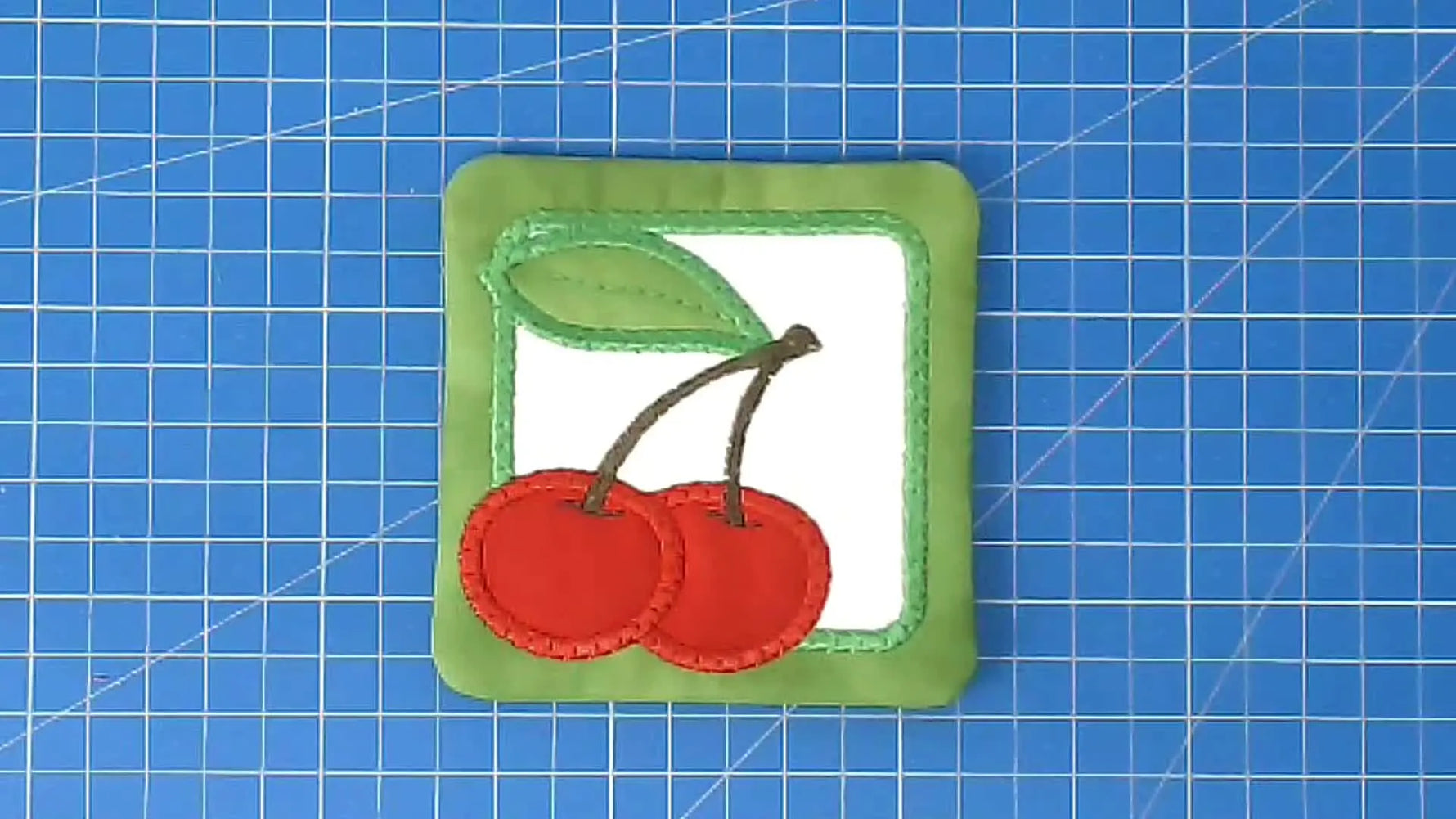 A finished in-the-hoop cherry coaster lies flat on a cutting mat, showing crisp satin borders and a clean, gift-ready finish.